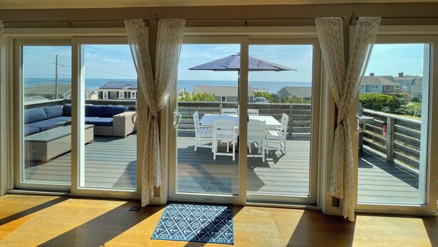 Una habitación con grandes puertas corredizas de vidrio que dan a un patio exterior con mesa y sillas, un sofá y vistas al océano y casas vecinas.
