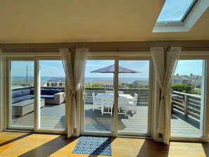 A room with large sliding glass doors leading to an outdoor patio area with a table and chairs, a sofa, and a view of the ocean and neighboring houses. - Gorgeous Beach Home Offering PANORAMIC Views Of Cape Cod Bay. (Sandwich)