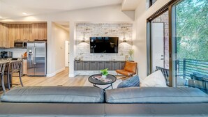 Cocina moderna con horno empotrado y microondas, comedor con mesa y sillas de madera, sala de estar con sofá y mesa de centro, y un ventanal con vistas a árboles.