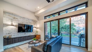 Una sala de estar moderna con pared de piedra, un televisor de pantalla plana, un sofá de cuero, una mesa de centro y un amplio ventanal corredero que da a un patio exterior.