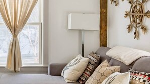 Een woonkamer met een grijze bank, gekleurde kussens, een houten salontafel, een lamp en een raam met gordijnen.