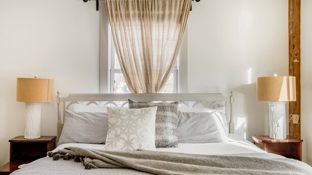 Een net opgemaakt bed met gestreept bedlinnen, een beige deken en patroon kussens. Twee bedlampen staan op houten nachtkastjes.