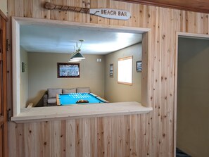 Une table de billard dans une pièce aux murs lambrés, une enseigne indiquant « Bar de plage », et des tableaux encadrés accrochés au mur.
