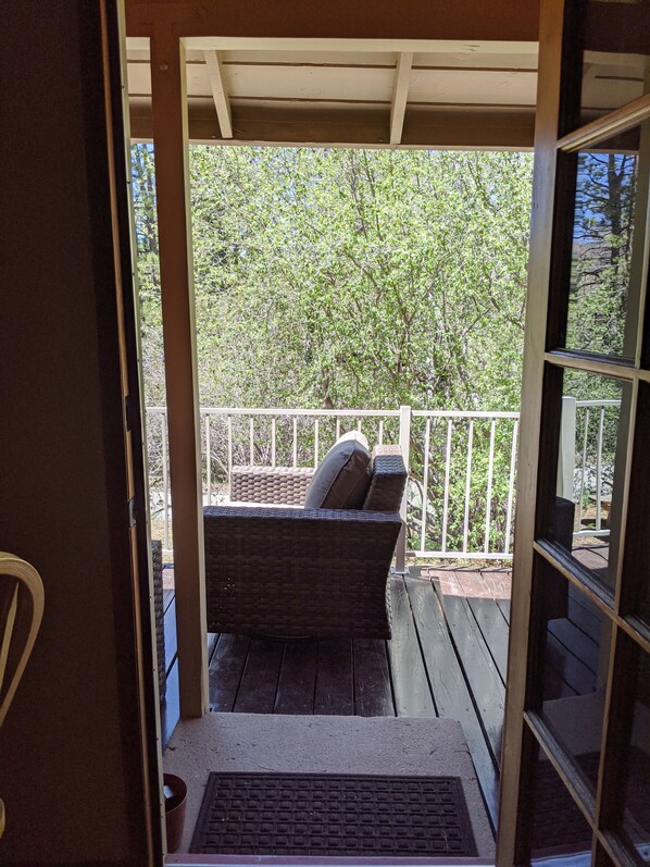 Une terrasse en bois avec un canapé en osier et des coussins, une rambarde en métal et une vue sur les arbres.