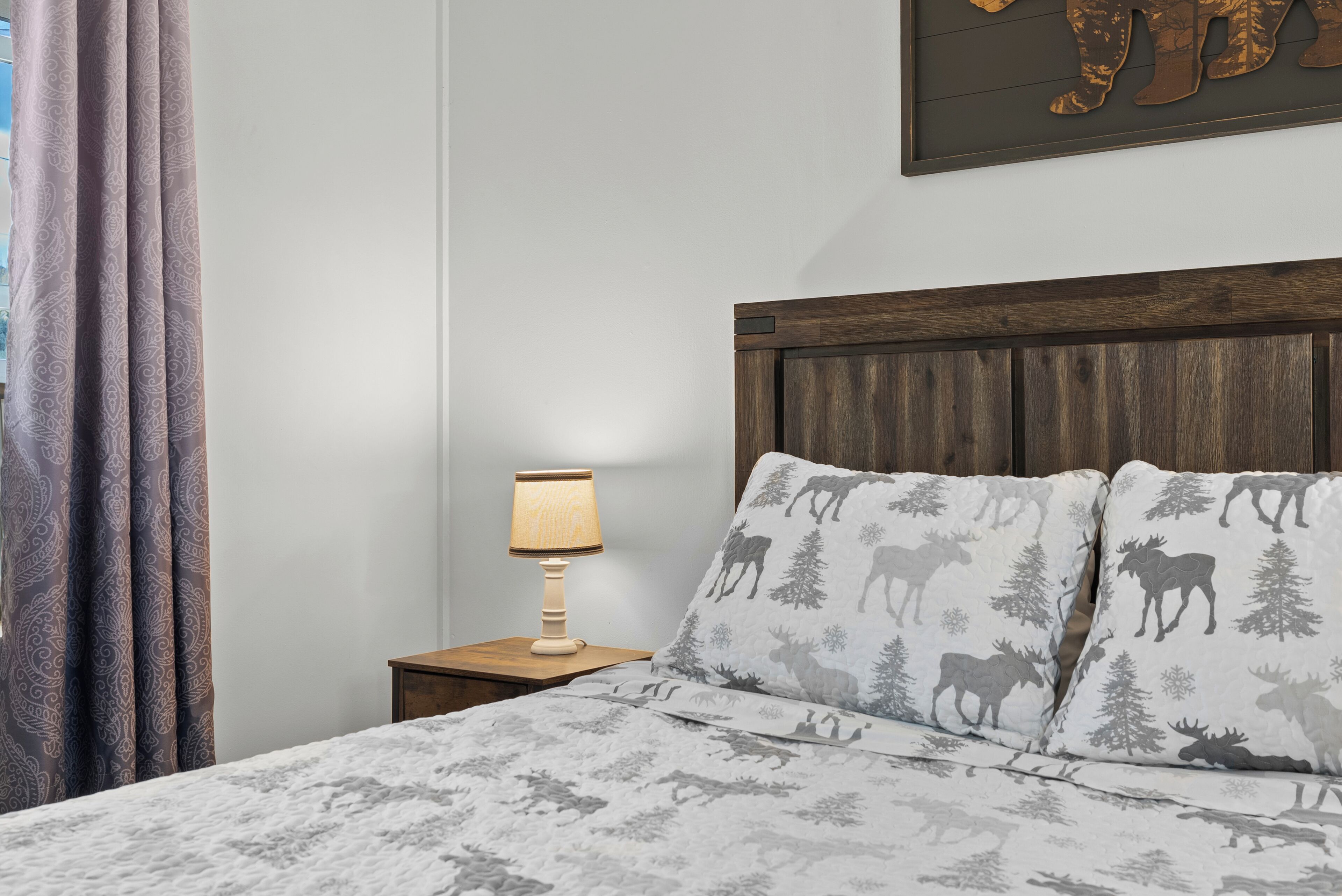 Ein Schlafzimmer mit einem Bett, zwei Kissen mit Elchmuster, einem Nachttisch mit Lampe und einem Wandbild.