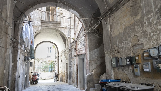 An arched corridor with a motorcycle parked at the end, electrical boxes on the walls, and a sign indicating non-recyclable waste. - Maiorani Family Apartment by Wonderful Italy (Napoli)