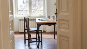 A room with a wooden chair, a table with a book and a cup, and a window with a view of buildings. - Maiorani Family Apartment by Wonderful Italy (Napoli)