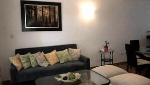 Ein Wohnzimmer mit einem dunklen Sofa, gemusterten Kissen, einem Glastisch und einem gerahmten Waldgemälde an der Wand.