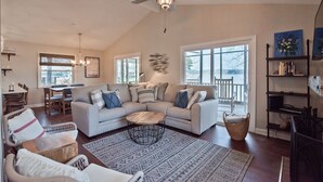 A spacious living room with a large sectional sofa, a coffee table, and a ceiling fan. There is a dining area with a table and chairs, and a bookshelf with books and decorative items. - Lake Tillery- Beautiful sunrises, large screened porch, and newly renovated home (Norwood)