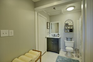 Un baño con inodoro blanco, mueble oscuro y un espejo sobre el lavamanos. Hay toallas en un perchero y una cortina de ducha.