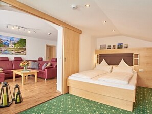 A bedroom with a bed, a wooden headboard, a green patterned rug, a wooden floor, a red sofa, a small table with a vase, and a painting on the wall.