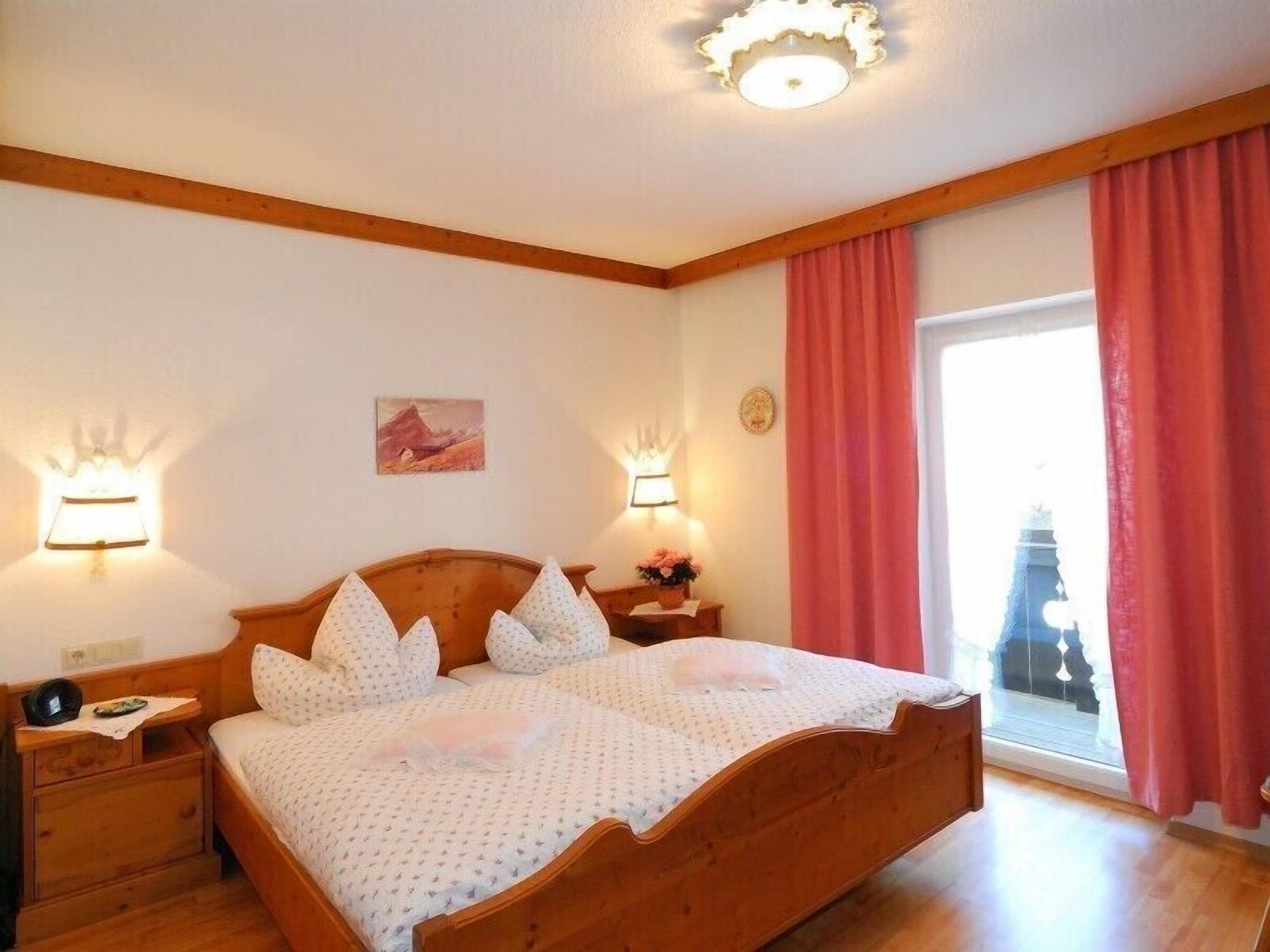 Une chambre à coucher avec un lit en bois, deux tables de chevet, une commode, une lampe fixée au mur, un tableau et une fenêtre avec des rideaux.