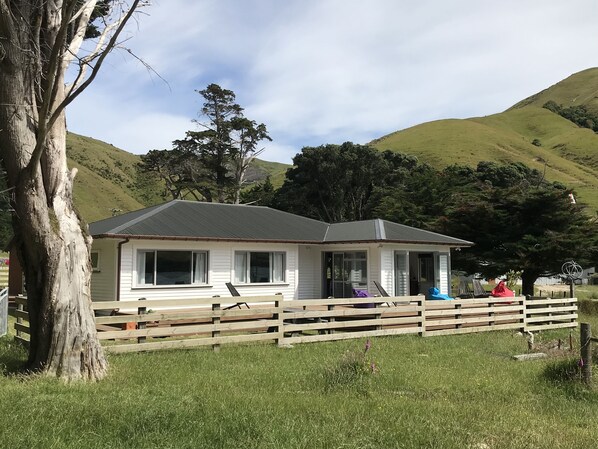 Ein weißes Einfamilienhaus mit grauem Dach, umgeben von einem Holzzaun, eingebettet in eine grüne Wiese mit Hügeln im Hintergrund.