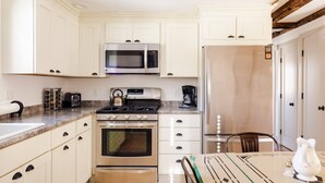 Une cuisine avec des armoires blanches, une cuisinière en acier inoxydable, un four à micro-ondes et un réfrigérateur. On y trouve une table recouverte d’un chemin de table vert et blanc, ainsi qu’un salier.