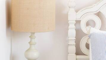Un lit blanc, richement décoré, avec une tête de lit incurvée, une table de chevet ronde en bois surmontée d’une lampe blanche, et un coussin à rayures.