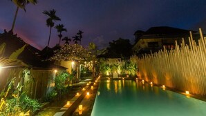 Vue nocturne d’un espace piscine avec une clôture en bois, des palmiers et un bâtiment en arrière-plan.