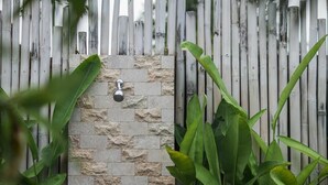 Un mur de pierre agrémenté d’un pommeau de douche en métal, entouré d’une clôture en bambou et de verdure.