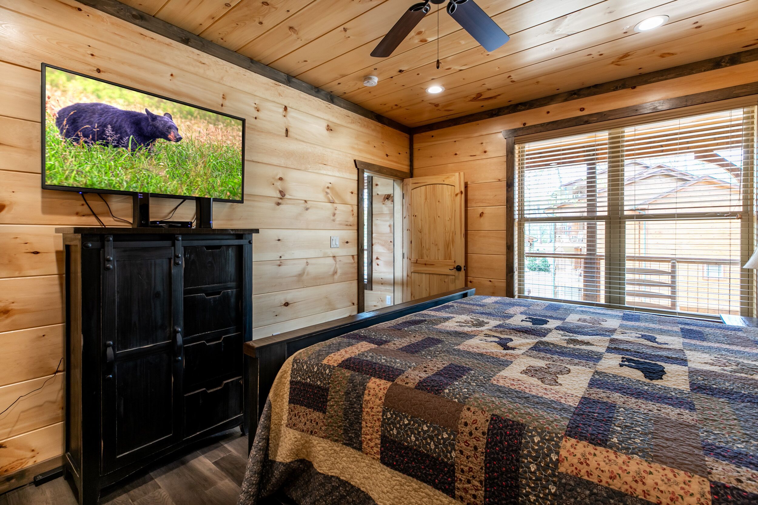 A bedroom with a wooden ceiling, a television mounted on the wall, a bed with a quilt, and a window with blinds.
