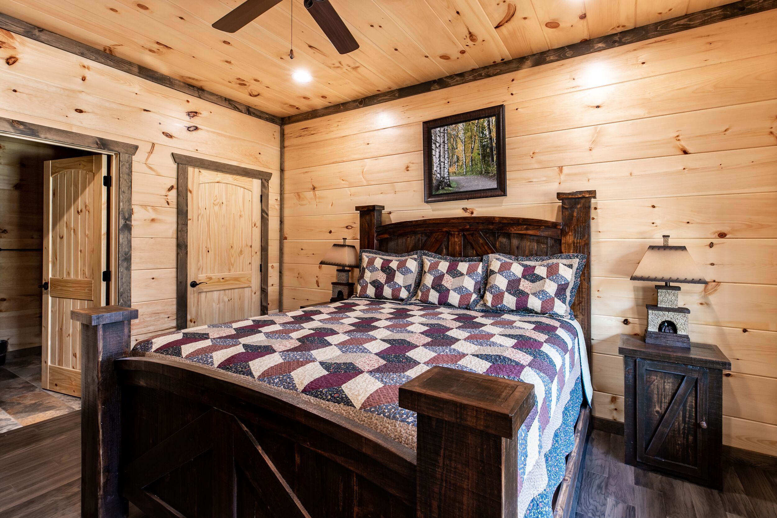 Un dormitorio de cabaña de madera con una cama, un ventilador de techo, una mesita de noche y un cuadro enmarcado en la pared.