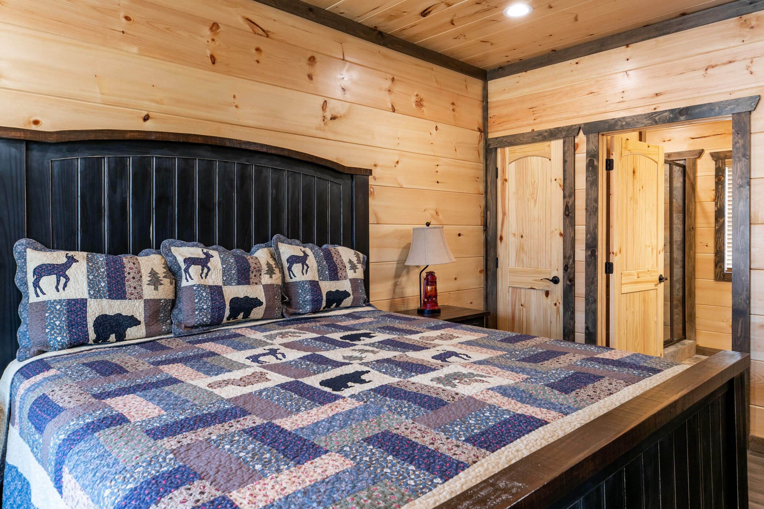 Une pièce aux murs lambrissés, avec un lit recouvert d’une courtepointe à motifs animaliers, une table de chevet avec une lampe et une porte dotée d’un cadre en bois.