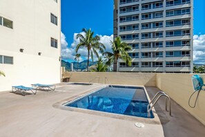 A rooftop pool area with two lounge chairs, a small hot tub, and a view of a tall building and palm trees.