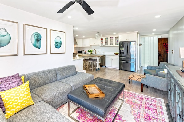 A modern living room with a grey sofa, a black ottoman, and a colorful rug. There is a kitchen in the background with white cabinets and a stainless steel refrigerator.