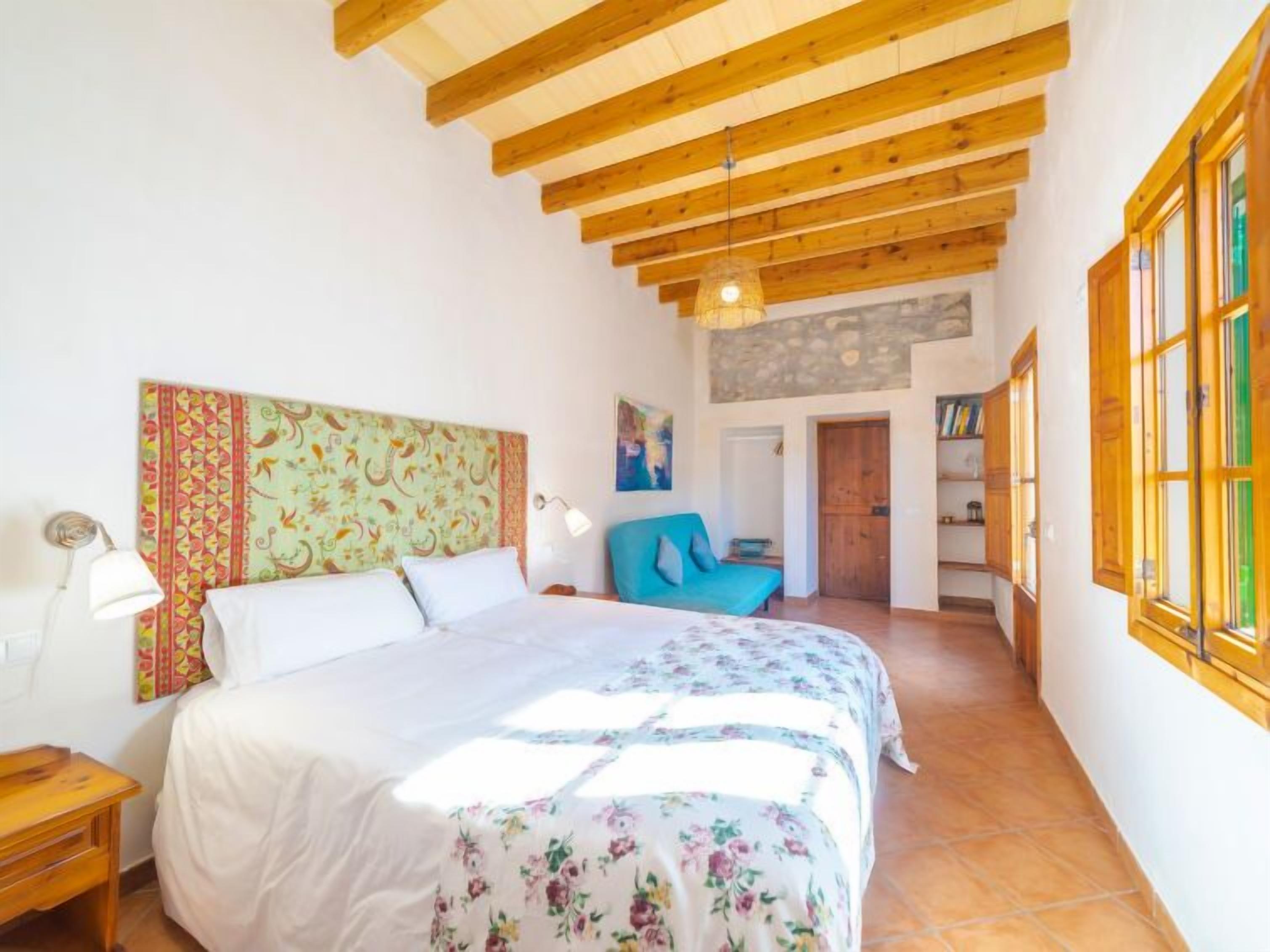 A bedroom with a wooden ceiling, a bed with floral bedding, a blue chair, and a bookshelf.