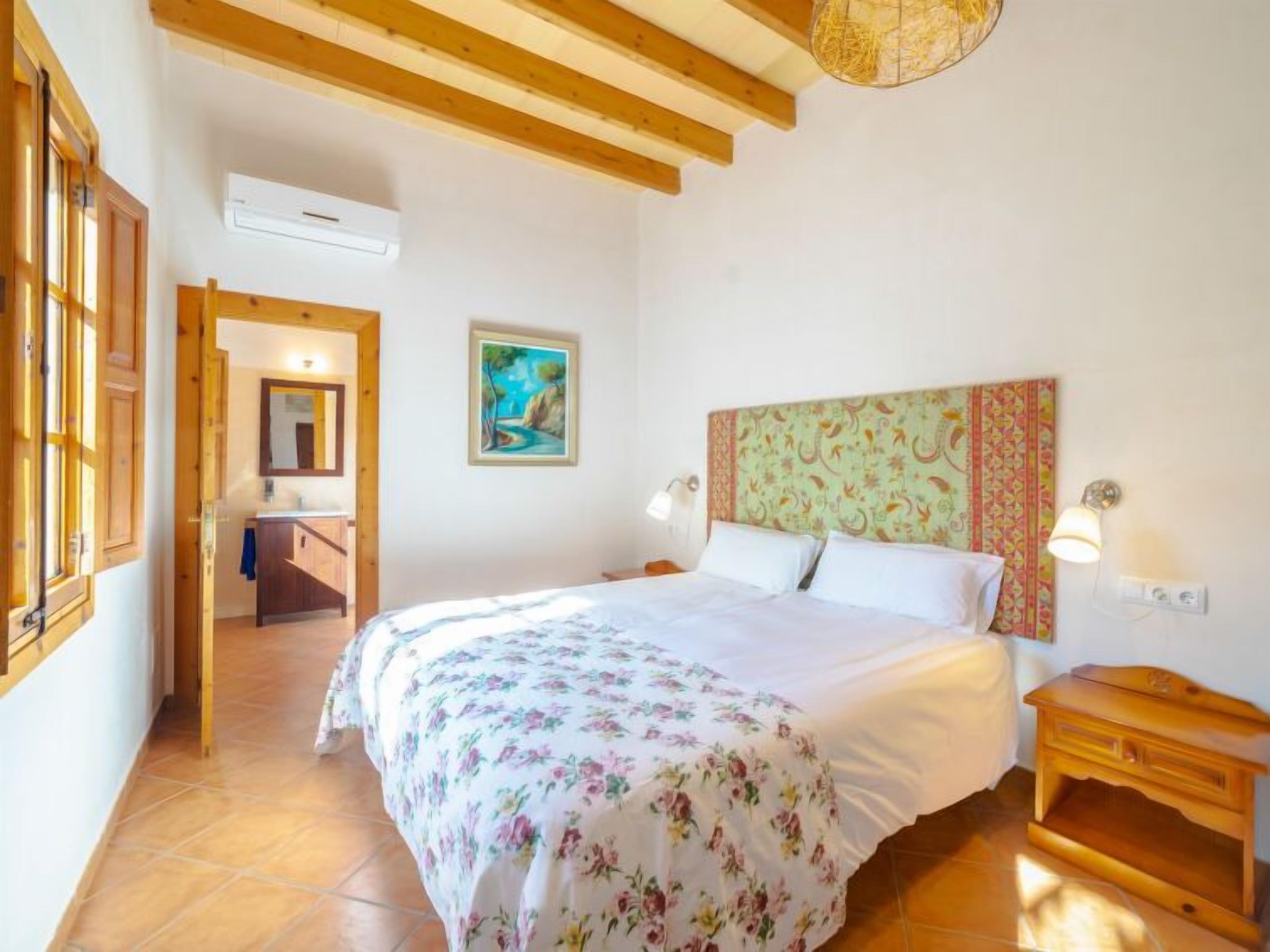 A bedroom with a bed, wooden headboard, a painting, a bedside table, and an air conditioning unit.