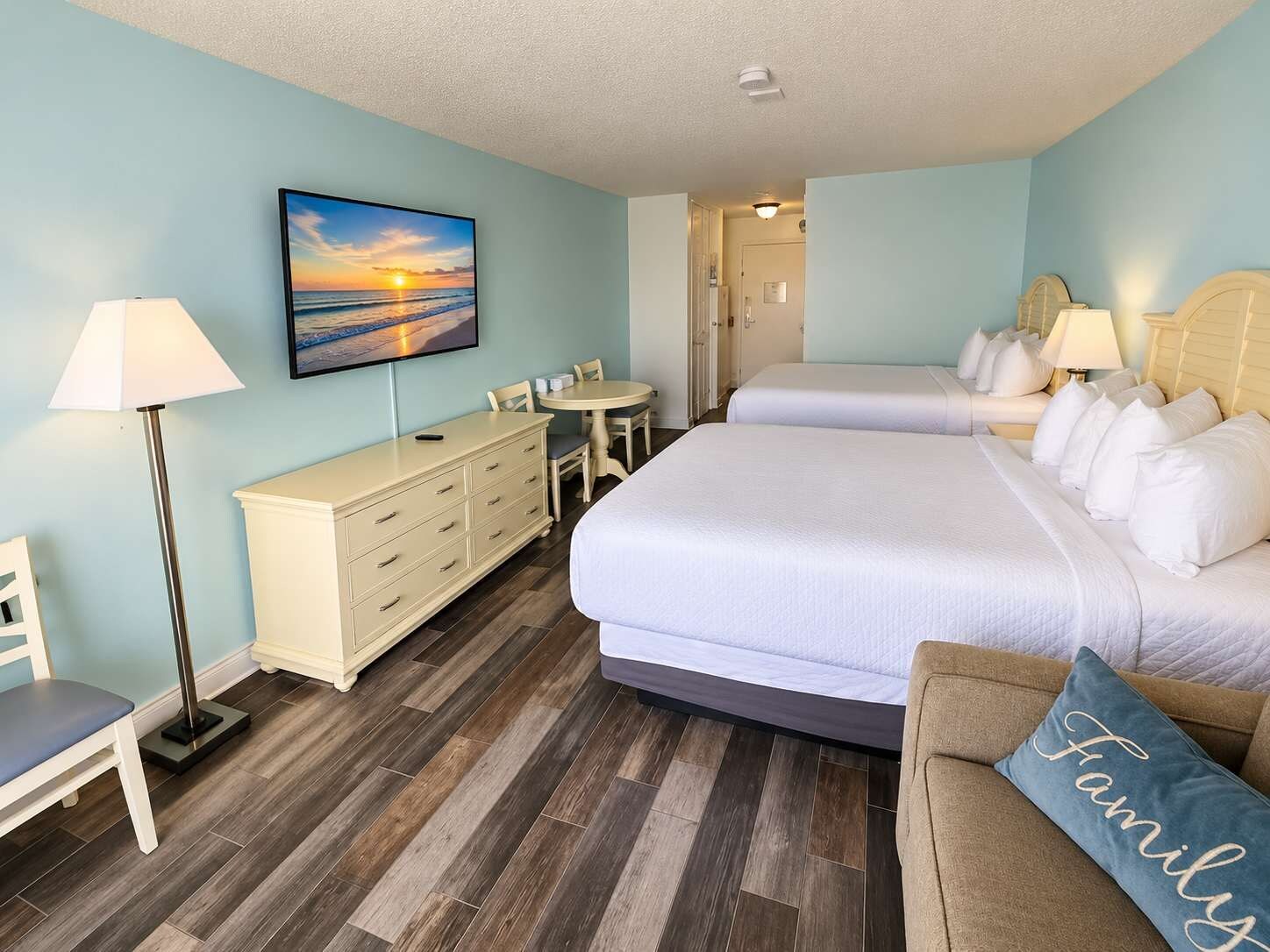 A hotel room with a bed, a desk, a chair, a lamp, and a painting of a sunset.