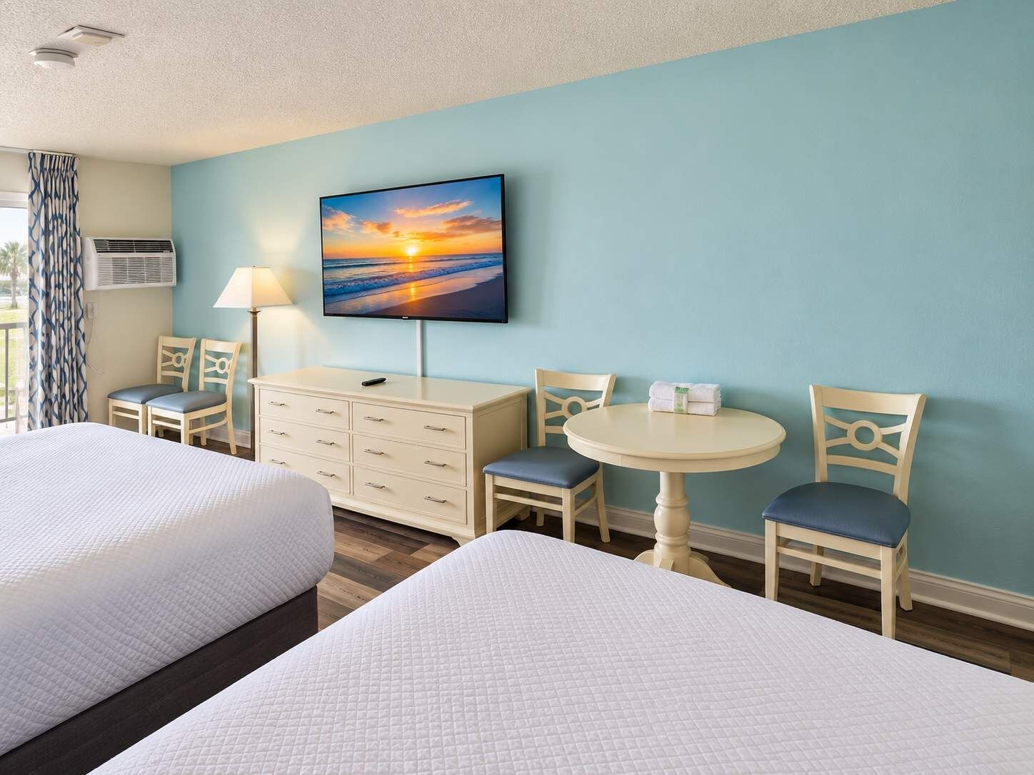 A hotel room with two beds, a desk, a chair, a table, a TV, and a window with curtains.