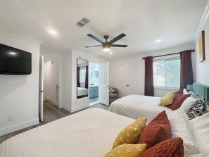 Two single beds with white bedding and decorative pillows, a ceiling fan, flat-screen TV, and a window with curtains.