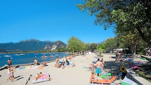 A beach with people sunbathing and swimming, boats in the water, and mountains in the background. - Lovely apartment for 6 guests with pool, WIFI, TV and balcony (Verbania)
