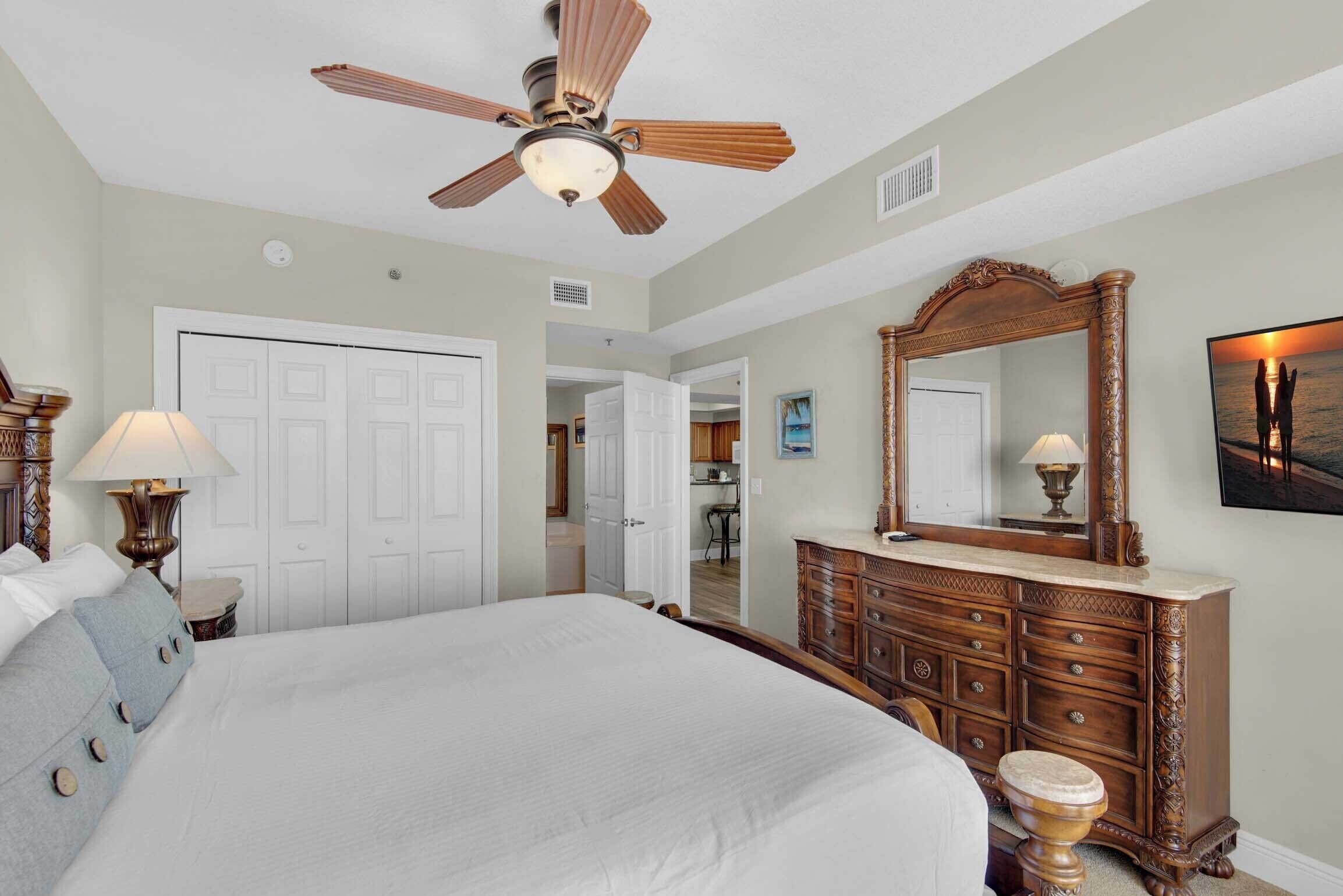A bedroom with a large bed, a wooden dresser, a ceiling fan, and a mirror.