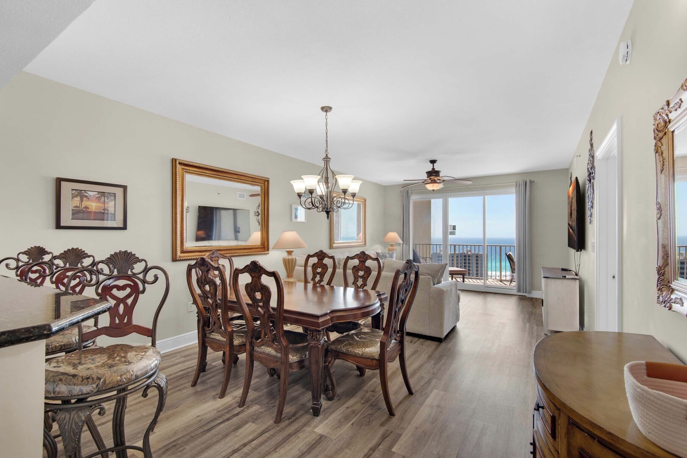 A dining area with a wooden table and chairs, a chandelier, and a view of the ocean through large windows.