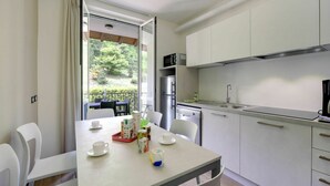 Eine moderne Küche mit weißen Schränken, einem Edelstahlkühlschrank und einem Geschirrspüler. Ein Esstisch mit Stühlen bietet Blick auf Grünflächen.