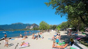 Una playa con gente tomando el sol y nadando, barcos en el agua y montañas al fondo.