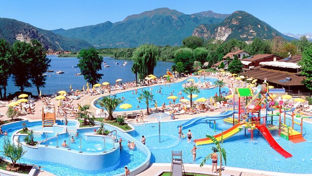 Una amplia piscina al aire libre con varias pistas y un tobogán acuático, rodeada de árboles y montañas.