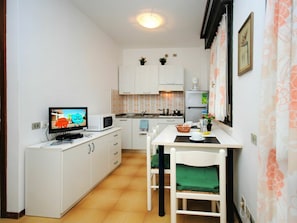 Cocina compacta con armarios blancos, un microondas, una pequeña mesa de comedor con sillas y un televisor mostrando una caricatura.