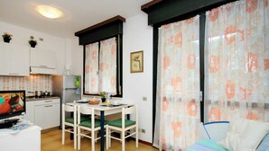 A compact room with a bed, a dining table with chairs, a small kitchenette, and a window with floral curtains.