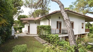 A two-story house with a balcony, surrounded by lush greenery and a small garden. - Beautiful apartment for 7 people with A/C, TV, terrace and pets allowed (Lignano Sabbiadoro)
