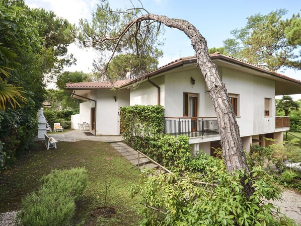 Una casa de dos pisos con balcón, rodeada de abundante vegetación y un pequeño jardín.