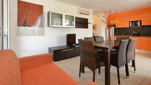 Ett modernt vardagsrum med en matplats, inrett med en soffa, ett matbord, stolar, en TV och ett kök med orange skåp.