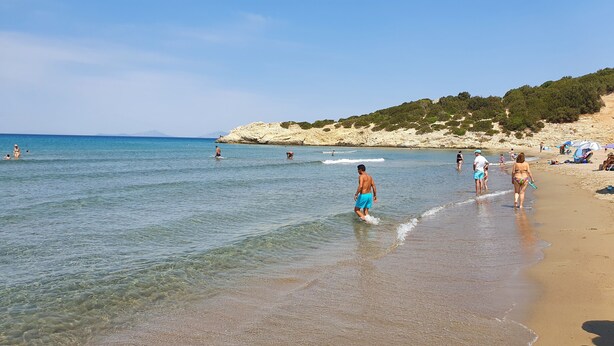 Una playa con agua cristalina, gente caminando y nadando, y una ladera rocosa al fondo.