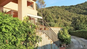 A two-story building with a staircase leading to a balcony, surrounded by lush greenery and a hillside in the background. - Beautiful apartment for 4 people with WIFI, TV, terrace and pets allowed (Rio Nell'elba)