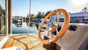 A boat's steering wheel with a wooden grip and a logo, a wooden deck, and a marina with boats in the background. - Awesome ship in Hennigsdorf with lake view (Hennigsdorf)