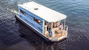 A houseboat with a deck, a small cabin, and a person sitting on the deck. - Awesome ship in Hennigsdorf with lake view (Hennigsdorf)