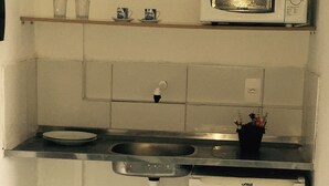 Une cuisine compacte équipée d’un four à micro-ondes, d’un petit réfrigérateur, d’un évier et d’une étagère où sont posées des tasses et un vase à fleurs.
