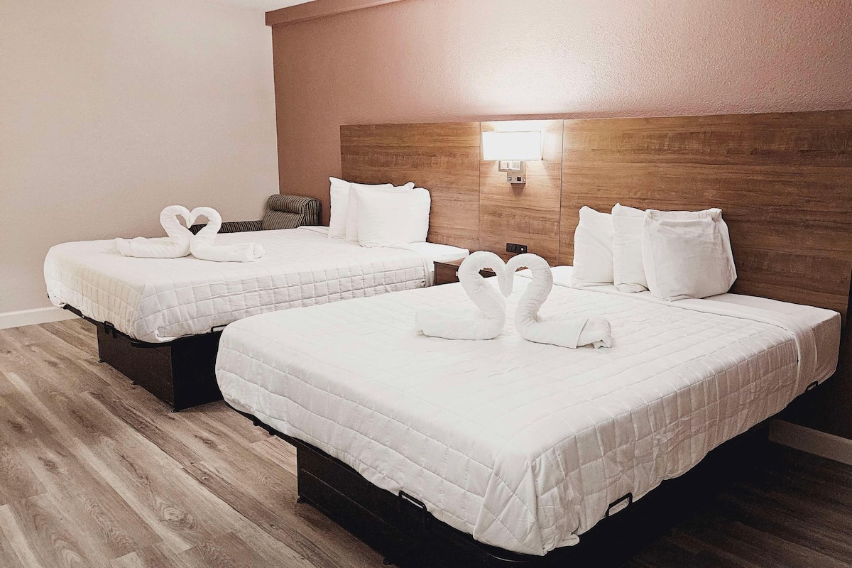 Duas camas com roupa de cama branca e toalhas em forma de cisne, uma cabeceira de madeira e uma luminária fixada na parede.