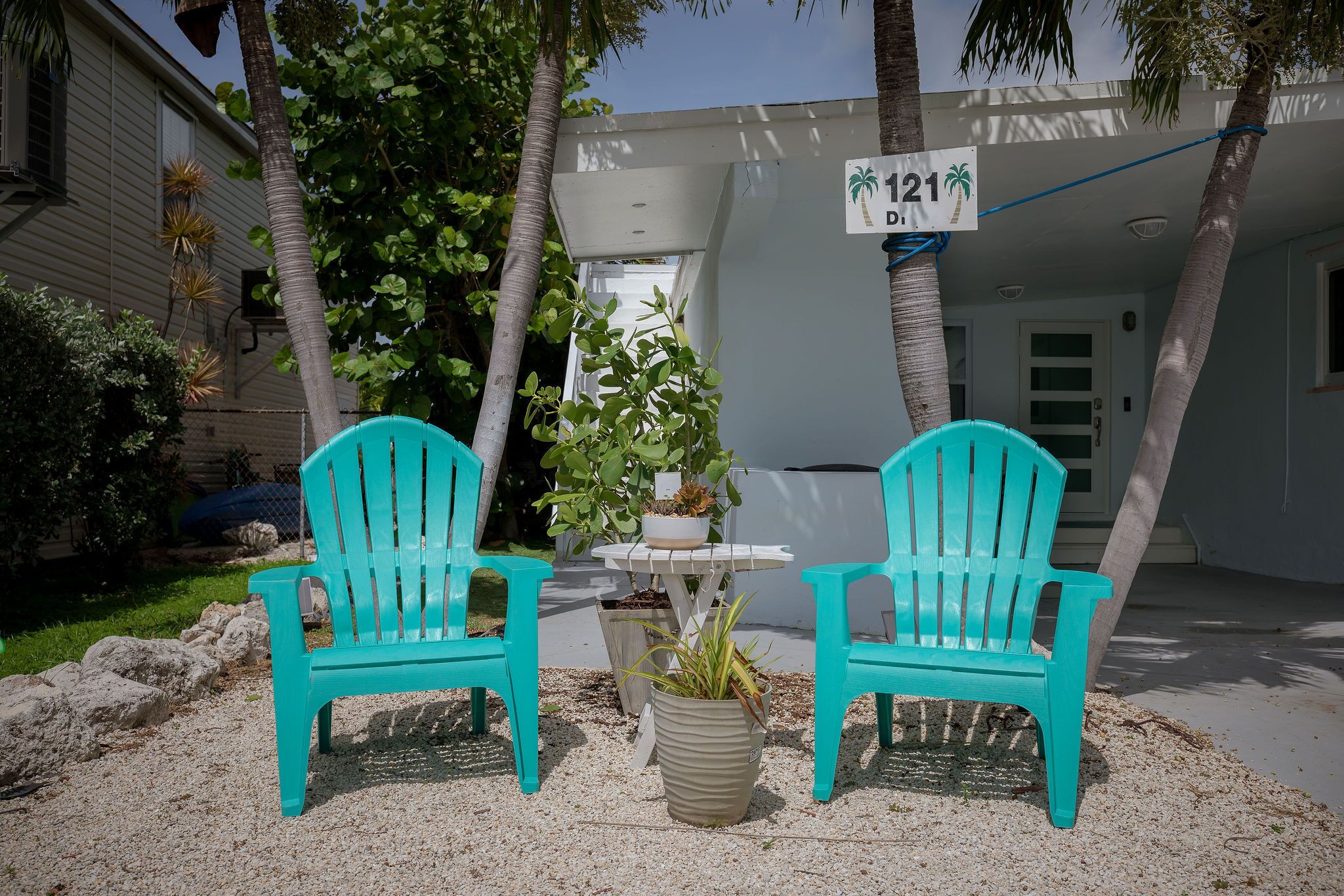 Deux chaises turquoise, une petite table et une plante en pot, le tout dans une cour avec un bâtiment en arrière-plan.