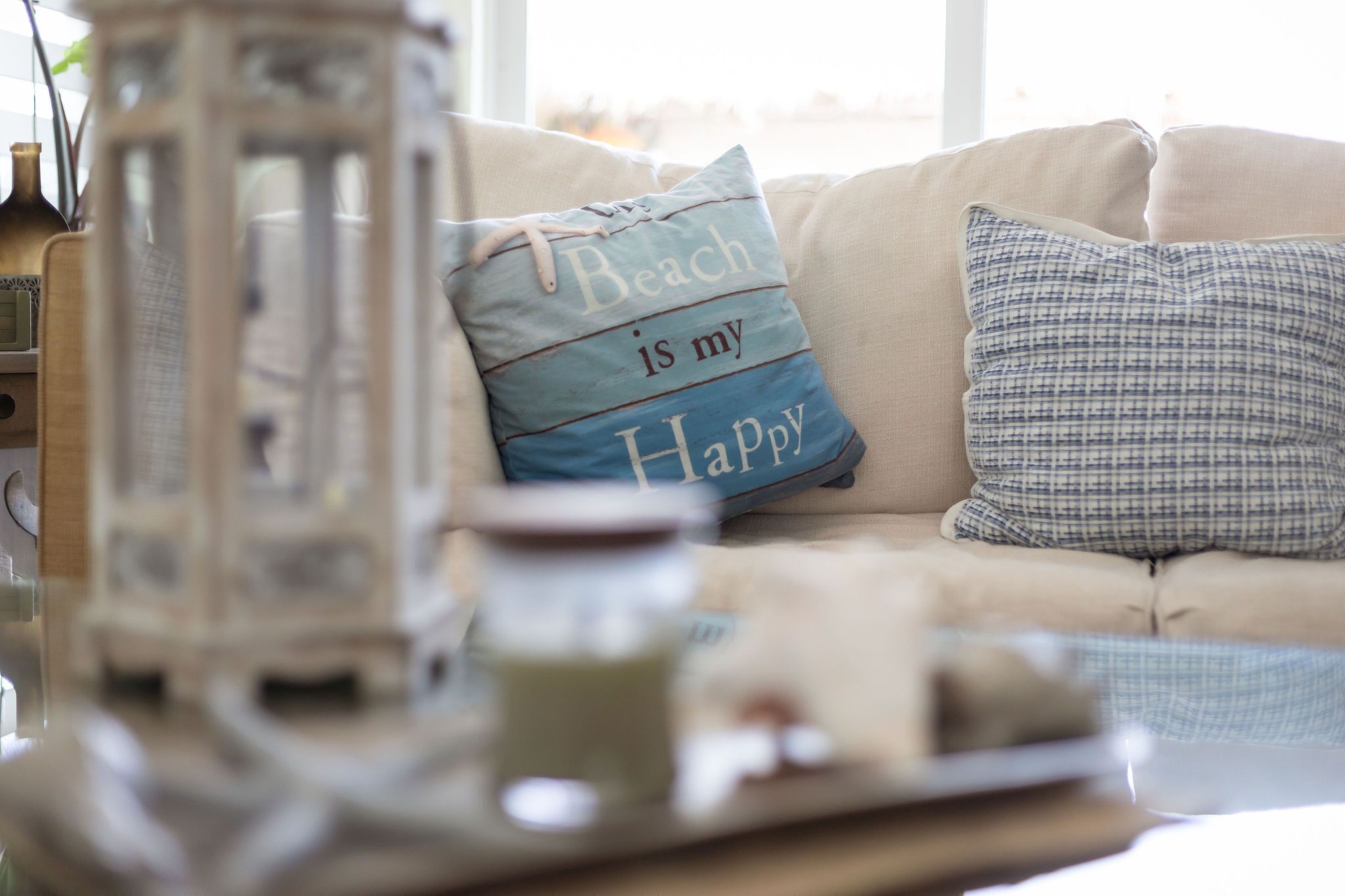 Ein gemütliches Wohnzimmer mit einem beigen Sofa, einem dekorativen Kissen mit dem Aufdruck "Beach is my Happy Place" und einer nostalgischen Laterne.